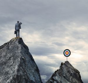 A businessman stands on a mountain top and looks towards a target on top of a distant mountain top in the distance. The businessman stands with his hand over his forehead as he peers towards the other peak wondering how he is going to get there.